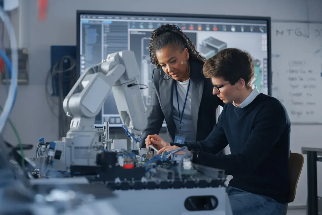 A imagem apresenta dois cientistas, uma mulher afro-americana jovem e um rapaz branco, ambos com roupas formais, trabalhando em frente a uma mesa com uma mão robótica. O tema do artigo é tecnologia e inovação.